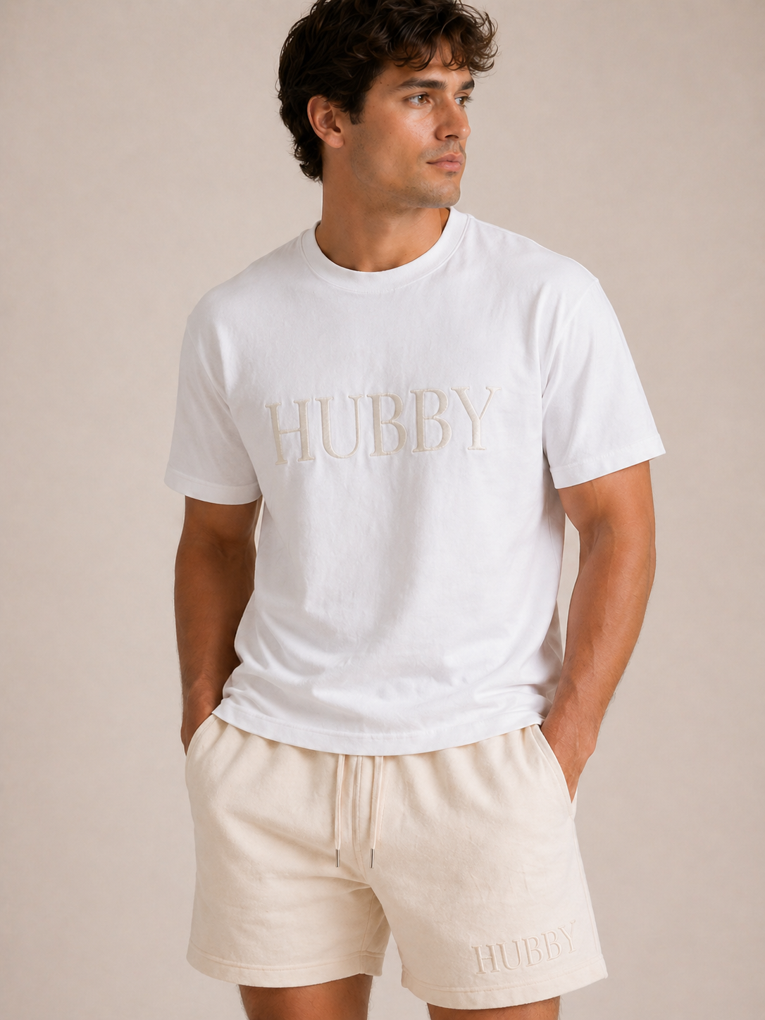 Man wearing a white t-shirt with 'HUBBY' text and beige shorts on a plain background