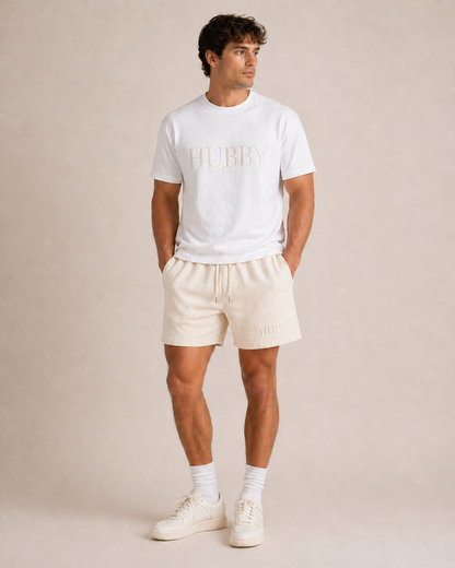 Man wearing a white 'HUBBY' t-shirt and beige shorts on a plain background